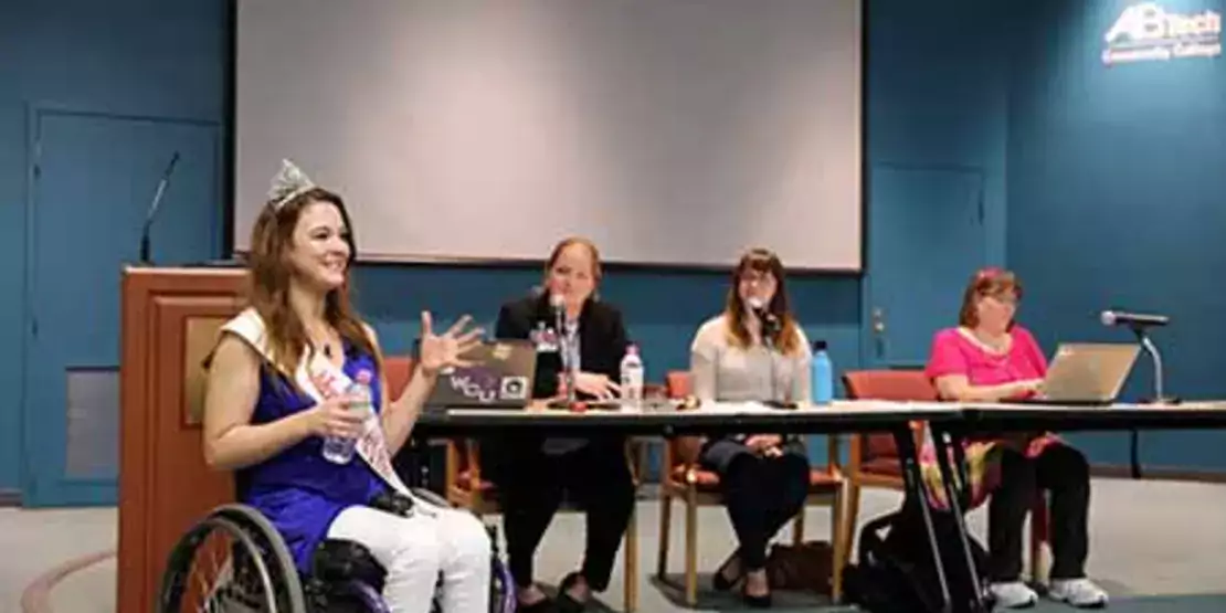 Madeline Delp in a wheelchair in front of three women at a table. 