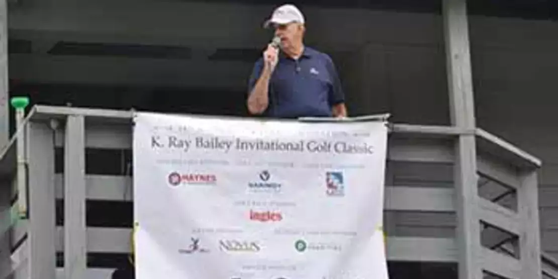 K. Ray Bailey speaking on deck with sign full of sponsors.